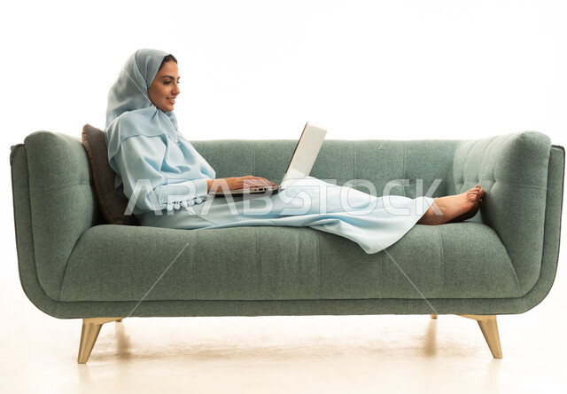 إنجاز المهام المطلوبة عبر اللابتوب، ادارة الأعمال عن بعد، استخدام الأجهزة الحديثة والمتطورة، بورتريه لامرأة عربية خليجية سعودية ترتدي العباءة والحجاب تجلس على الاريكة تستخدم الحاسب المحمول ، دمج التكنولوجيا بالعمل، مهن ووظائف نسائية، خلفية بيضاء