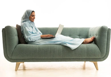 Completing required tasks via laptop, managing business remotely, using modern and advanced devices, a portrait of a Saudi Gulf Arab woman wearing an abaya and hijab sitting on a sofa using a laptop, integrating technology with work, women's professions and jobs, white background.