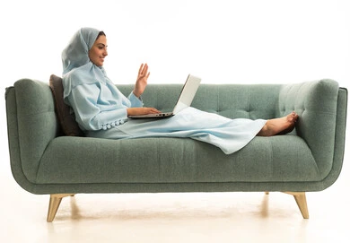 اجراء مكالمة فيديو عبر الانترنت، رفع كف اليد بتعابير التحية، إنجاز المهام المطلوبة عبر اللابتوب، ادارة الأعمال عن بعد، استخدام الأجهزة الحديثة والمتطورة، بورتريه لامرأة عربية خليجية سعودية ترتدي العباءة والحجاب تجلس على الاريكة تستخدم الحاسب المحمول ، خلفية بيضاء