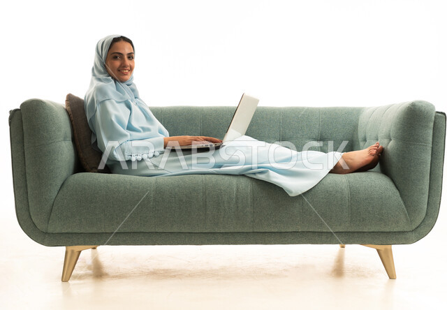 ادارة الأعمال عن بعد، استخدام الأجهزة الحديثة والمتطورة، بورتريه لامرأة عربية خليجية سعودية ترتدي العباءة والحجاب تجلس على الاريكة تستخدم الحاسب المحمول ، دمج التكنولوجيا بالعمل، إنجاز المهام المطلوبة عبر اللابتوب، مهن ووظائف نسائية، خلفية بيضاء