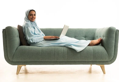 Remote business management, using modern and advanced devices, a portrait of an Arab Gulf Saudi woman wearing an abaya and hijab sitting on a sofa using a laptop, integrating technology with work, completing required tasks via the laptop, women's professions and jobs, white background.
