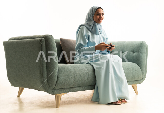 التقنيات الالكترونية الحديثة, مفهوم الاندماج والتفاعل باللعب في ألعاب البلايستيشن الترفيهية، بورتريه لإمرأة عربية سعودية خليجية تجلس على الاريكة تحمل بيدها عصا التحكم، قضاء إجازة ممتعة, خلفية بيضاء