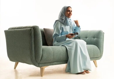 Spending free time in video games, gestures and facial expressions indicating victory and triumph, a portrait of a Saudi Arabian Gulf woman wearing a hijab and abaya sitting on the couch holding a game controller and playing video games, white background.