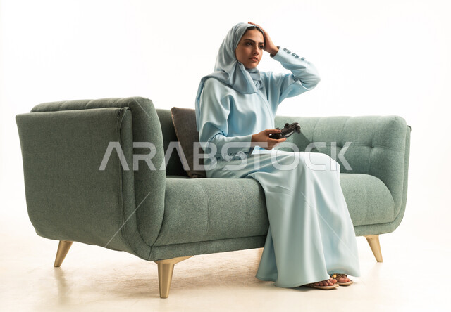 Expressions of sadness and loss, the use of technological devices and modern techniques, a portrait of a young Arab Gulf Saudi woman wearing an abaya and hijab sitting on the sofa holding a game controller with her hand on her head in gestures of regret, playing recreational video games, white background.