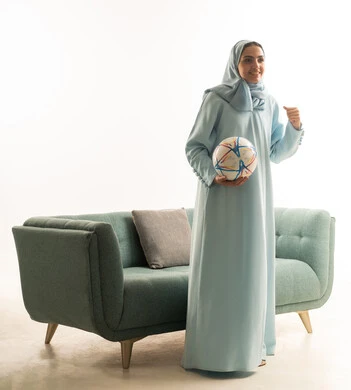 تشجيع الفريق المفضل بحماس ومتعة، الاهتمام بالرياضة ومتابعة المباريات والدوريات المختلفة، بورتريه لشابة عربية خليجية سعودية ترتدي العباءة و الحجاب تقف بجانب الاريكة تحمل الكرة وترفع يدها بإيماءات الاندماج والسعادة بفوز ناديها، الجسم بالطول الكامل، خلفية بيضاء