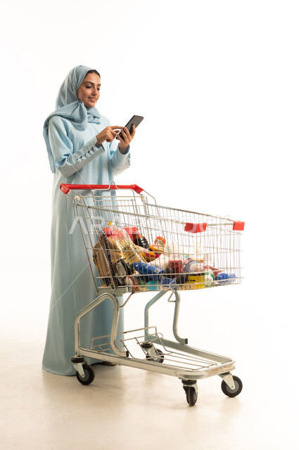 تصفح مواقع التواصل الاجتماعي عبر الجوال، انتقاء الأفضل والأجود من السلع الموجودة في السوبر ماركت بالمملكة العربية السعودية، إمرأة عربية خليجية سعودية مبتسمة ترتدي العباءة والحجاب تمسك بعربة التسوق، الاستفادة من العروض والتخفيضات، الجسم بالطول الكامل، خلفية بيضاء