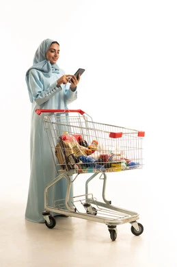 تصفح مواقع التواصل الاجتماعي عبر الجوال، انتقاء الأفضل والأجود من السلع الموجودة في السوبر ماركت بالمملكة العربية السعودية، إمرأة عربية خليجية سعودية مبتسمة ترتدي العباءة والحجاب تمسك بعربة التسوق، الاستفادة من العروض والتخفيضات، الجسم بالطول الكامل، خلفية بيضاء
