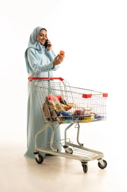 اجراء مكالمة هاتفية عبر الجوال، انتقاء الأفضل والأجود من السلع الموجودة في السوبر ماركت بالمملكة العربية السعودية، إمرأة عربية خليجية سعودية مبتسمة ترتدي العباءة والحجاب تقف امام عربة التسوق تمسك بيدها نوع من المنتجات، الاستفادة من العروض والتخفيضات، الجسم بالطول الكامل، خلفية بيضاء
