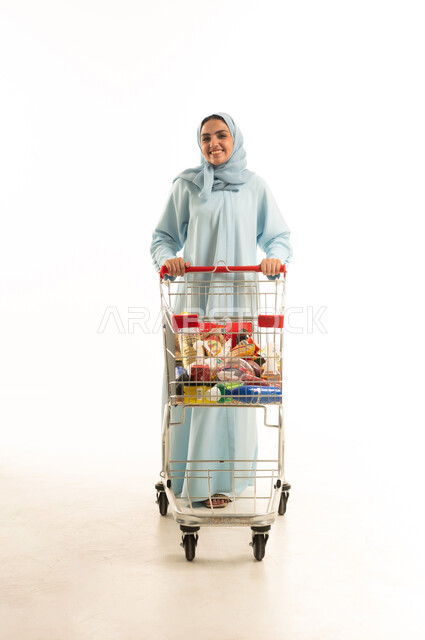 الاستمتاع بالتسوق في المولات والمراكز التجارية، شراء المستلزمات والحاجيات الضرورية، الاستفادة من الخصومات على المنتجات، بورتريه لامرأة عربية خليجية سعودية محجبة ترتدي العباءة التقليدية وتجر عربة التسوق أمامها بإيماءات السعادة، الجسم بالطول الكامل، خلفية بيضاء