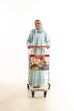 الاستمتاع بالتسوق في المولات والمراكز التجارية، شراء المستلزمات والحاجيات الضرورية، الاستفادة من الخصومات على المنتجات، بورتريه لامرأة عربية خليجية سعودية محجبة ترتدي العباءة التقليدية وتجر عربة التسوق أمامها بإيماءات السعادة، الجسم بالطول الكامل، خلفية بيضاء