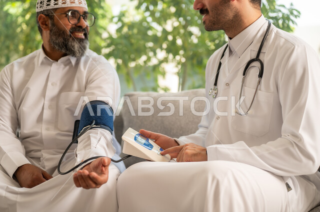 العمل في القطاع الصحي، الإصابة بوعكة صحية، استخدام الميزان الاكتروني الحديث والمتطور للفحص المنزلي السريع الحديث، الخدمات الطبية والرعاية الصحية، ممرض يرتدي المعطف الابيض يقوم بقياس ضغط الدم لرجل مسن عربي خليجي سعودي يلبس الثوب التقليدي يجلس في المنزل