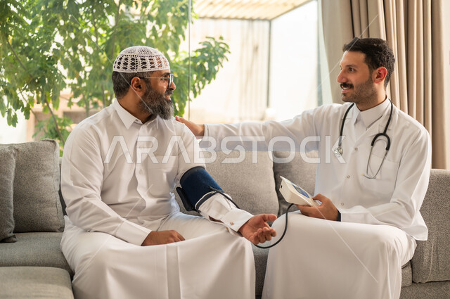 استخدام الميزان الاكتروني الحديث والمتطور للفحص المنزلي السريع الحديث، الخدمات الطبية والرعاية الصحية، ممرض يرتدي المعطف الابيض يقوم بقياس ضغط الدم لرجل مسن عربي خليجي سعودي يلبس الثوب التقليدي يجلس في المنزل، الإصابة بوعكة صحية، العمل في القطاع الصحي