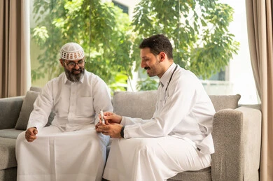العناية الصحية والفحص الطبي الفوري، جهاز قياس سكر الدم لمرضى السكري، العمل في القطاع الطبي وشركات التمريض المنزلي, ممرض يرتدي المعطف الطبي يقوم بفحص نسبة السكر في الدم لمريض مسن عربي خليجي سعودي يرتدي الثوب التقليدي و القبعة يجلس على الكنبه