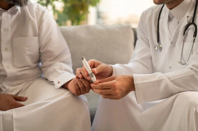 توفير خدمات الرعاية الصحية والفحص الطبي السريع، جهاز قياس سكر الدم لمرضى السكري، العمل في القطاع الطبي وشركات التمريض المنزلي,  صورة مقربة لممرض يرتدي المعطف الطبي يقوم بفحص نسبة السكر في الدم لمريض مسن عربي خليجي سعودي يرتدي الثوب التقليدي و القبعة يجلس على الكنبه