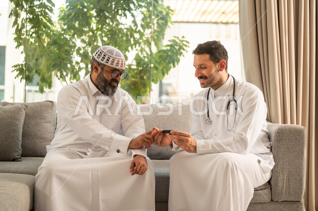 العمل في القطاع الطبي وشركات التمريض المنزلي، جهاز قياس سكر الدم لمرضى السكري، الرعاية الصحية والفحص الطبي السريع, ممرض يرتدي المعطف الطبي يقوم بفحص نسبة السكر في الدم لمريض مسن عربي خليجي سعودي يرتدي الثوب و القبعة البيضاء يجلس على الكنبه