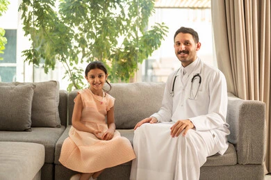 شركات التمريض المنزلي في السعودية، الرعاية الطبية للأطفال, مفهوم الاهتمام بالصحة الأسرية, طبيب عربي خليجي سعودي يرتدي المعطف الطبي يجلس مع طفلة داخل المنزل، النظر للكاميرا بتعابير السرور