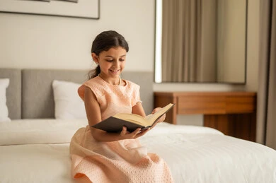 الاهتمام بتصفح الكتب المفيدة، تنمية مهارات القراءة للأطفال، مطالعة القصص التصويرية المسلية، تمضية أوقات الفراغ بالتعلم والتثقف الذاتي،  فتاة عربية خليجية سعودية ترتدي الفستان تجلس على السرير تمسك بيديها كتاب