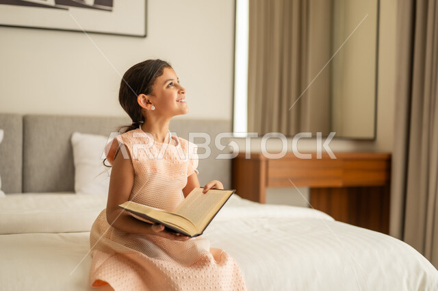 مطالعة القصص التصويرية المسلية، تمضية أوقات الفراغ بالتعلم والتثقف الذاتي،  فتاة عربية خليجية سعودية ترتدي الفستان تجلس على السرير تمسك بيديها كتاب, تنمية مهارات القراءة للأطفال, الاهتمام بتصفح الكتب المفيدة