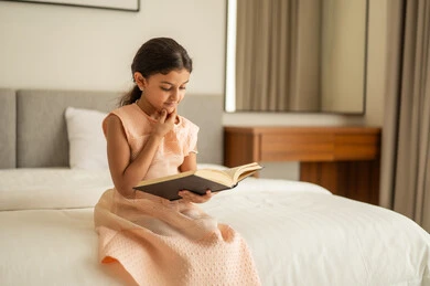 تمضية أوقات الفراغ بالتعلم والتثقف الذاتي، إيماءات التأمل والتمعن،  فتاة عربية خليجية سعودية ترتدي الفستان تجلس على السرير تمسك بيديها كتاب تضع يدها على ذقنها،  مطالعة القصص التصويرية المسلية, تنمية مهارات القراءة للأطفال, الاهتمام بتصفح الكتب المفيدة