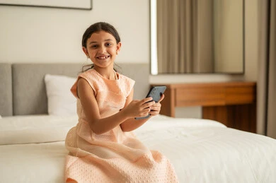 متابعة الدروس عن بعد عن طريق الانترنت، الدردشة والتواصل مع الاهل والأصدقاء، تصفح مواقع التواصل الاجتماعي، فتاة عربية خليجية سعودية ترتدي الفستان تجلس على السرير تحمل بيدها الهاتف المحمول، استخدام جهاز تقني حديث ومتطور، ايماءات السعادة و السرور