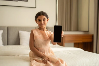 A blank black screen displayed on the mobile phone, browsing social media sites, a Gulf Arab Saudi girl wearing a dress sitting on the bed holding a mobile phone in her hand, using a modern and advanced technological device.