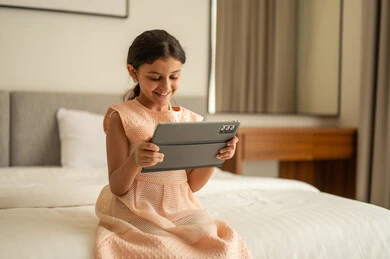 Monitoring available online e-learning platforms, a Gulf Arab Saudi girl wearing a dress sitting on the bed using a tablet, making a video call, integrating learning with technology and tech, watching recreational and educational activities.