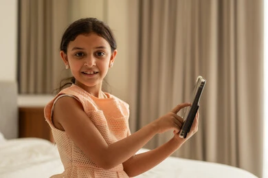 Making a video call, a Gulf Arab Saudi girl wearing a dress sitting on the bed using a tablet, integrating learning with technology and tech, following available online educational platforms, watching recreational and educational activities.