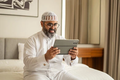 دمج العمل مع التكنولوجيا، إدارة شؤون الأعمال وتنظيمها عبر الاجهزة التقنية، العمل عن بعد وانجاز المهام المطلوبة، مهنة ووظيفة ادارية، رجل عربي خليجي سعودي يرتدي الثوب التقليدي والقبعة البيضاء يجلس على السرير يعمل على الجهاز اللوحي، استخدام جهاز تقني حديث ومتطور