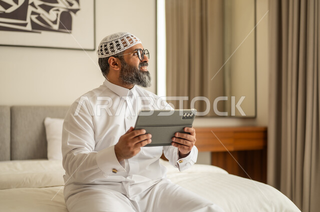 Looking at something, using a modern and advanced technical device, integrating work with technology, managing and organizing business affairs through technical devices, working remotely and completing required tasks, a profession and administrative job, an Arab Gulf Saudi man wearing traditional attire and a white cap sitting on the bed working on a tablet.