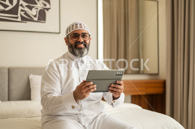 استخدام جهاز تقني حديث ومتطور، النظر للكاميرا بتعابير السرور، دمج العمل مع التكنولوجيا، إدارة شؤون الأعمال وتنظيمها عبر الاجهزة التقنية، العمل عن بعد وانجاز المهام المطلوبة، مهنة ووظيفة ادارية، رجل عربي خليجي سعودي يرتدي الثوب التقليدي والقبعة البيضاء يجلس على السرير يعمل على الجهاز اللوحي
