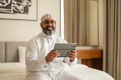 استخدام جهاز تقني حديث ومتطور، النظر للكاميرا بتعابير السرور، دمج العمل مع التكنولوجيا، إدارة شؤون الأعمال وتنظيمها عبر الاجهزة التقنية، العمل عن بعد وانجاز المهام المطلوبة، مهنة ووظيفة ادارية، رجل عربي خليجي سعودي يرتدي الثوب التقليدي والقبعة البيضاء يجلس على السرير يعمل على الجهاز اللوحي