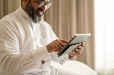 إدارة شؤون الأعمال وتنظيمها عبر الاجهزة التقنية، العمل عن بعد وانجاز المهام المطلوبة، مهنة ووظيفة ادارية، دمج العمل مع التكنولوجيا، رجل عربي خليجي سعودي يرتدي الثوب التقليدي والقبعة البيضاء يجلس على السرير يعمل على الجهاز اللوحي، استخدام جهاز تقني حديث ومتطور