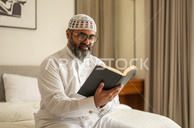 مفهوم التثقيف والبحث العلمي، رجل عربي خليجي سعودي يرتدي الثوب التقليدي والقبعة البيضاء يجلس على السرير يمسك كتاب، هواية القراءة والاستكشاف