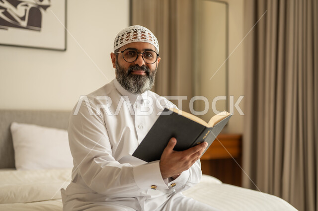 النظر للكاميرا بتعابير السرور، مفهوم التثقيف والبحث العلمي، رجل عربي خليجي سعودي يرتدي الثوب التقليدي والقبعة البيضاء يجلس على السرير يمسك بيدية كتاب، هواية القراءة والاستكشاف