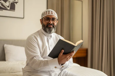 النظر للكاميرا بتعابير السرور، مفهوم التثقيف والبحث العلمي، رجل عربي خليجي سعودي يرتدي الثوب التقليدي والقبعة البيضاء يجلس على السرير يمسك بيدية كتاب، هواية القراءة والاستكشاف