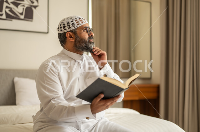 رجل عربي خليجي سعودي يرتدي الثوب التقليدي والقبعة البيضاء يجلس على السرير يمسك كتاب ويضع يده على ذقنه بإيماءات التفكير بشيء ما، قضاء وقت الفراغ بالتعلم والثقافة، مفهوم التثقيف والبحث العلمي، هواية المطالعة والاستكشاف