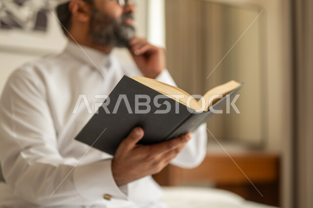 قضاء وقت الفراغ بالتعلم والثقافة، مفهوم التثقيف والبحث العلمي، هواية المطالعة والاستكشاف، رجل عربي خليجي سعودي يرتدي الثوب التقليدي والقبعة البيضاء يجلس على السرير يمسك كتاب ويضع يده على ذقنه بإيماءات التفكير بشيء ما