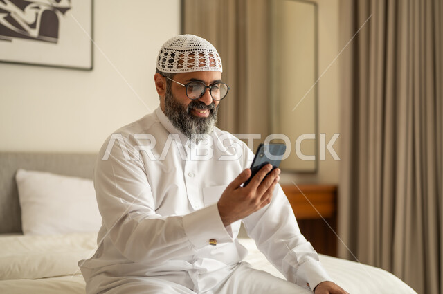 استخدام جهاز تقني حديث ومتطور، تصفح مواقع التواصل الاجتماعي، رجل عربي خليجي سعودي يرتدي الثوب التقليدي والقبعة البيضاء يجلس على السرير يمسك بيده الهاتف المحمول، الدردشة والتواصل مع الأهل و الأصدقاء، العمل عن بعد وانجاز المهام المطلوبة