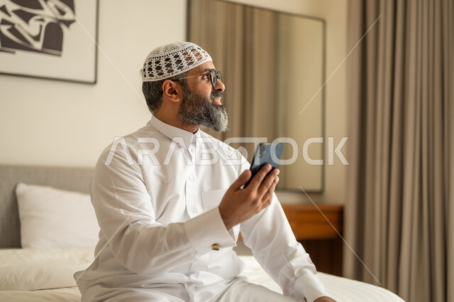 النظر الى شيء ما، تصفح مواقع التواصل الاجتماعي، رجل عربي خليجي سعودي يرتدي الثوب التقليدي والقبعة البيضاء يجلس على السرير يمسك بيده الهاتف المحمول، الدردشة والتواصل مع الأهل و الأصدقاء، استخدام جهاز تقني حديث ومتطور، العمل عن بعد وانجاز المهام المطلوبة