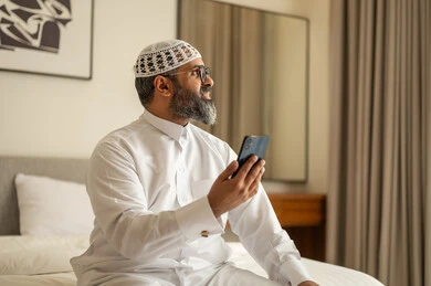 النظر الى شيء ما، تصفح مواقع التواصل الاجتماعي، رجل عربي خليجي سعودي يرتدي الثوب التقليدي والقبعة البيضاء يجلس على السرير يمسك بيده الهاتف المحمول، الدردشة والتواصل مع الأهل و الأصدقاء، استخدام جهاز تقني حديث ومتطور، العمل عن بعد وانجاز المهام المطلوبة