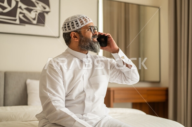 اجراء مكالمة هاتفية ، رجل عربي خليجي سعودي يرتدي الثوب التقليدي والقبعة البيضاء يجلس على السرير يمسك بيده الهاتف المحمول، الدردشة والتواصل مع الأهل و الأصدقاء، استخدام جهاز تقني حديث ومتطور، العمل عن بعد وانجاز المهام المطلوبة