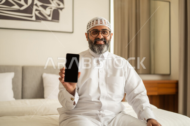 A blank black screen is displayed, browsing social media, a Saudi Arabian Gulf man wearing traditional attire and a white cap sits on the bed holding a mobile phone, using a modern and advanced technological device.