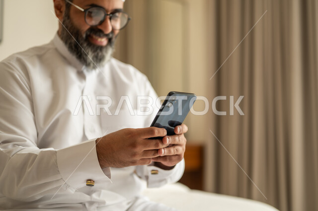 تصفح مواقع التواصل الاجتماعي، رجل عربي خليجي سعودي يرتدي الثوب التقليدي والقبعة البيضاء يجلس على السرير يمسك بيده الهاتف المحمول، الدردشة والتواصل مع الأهل و الأصدقاء، استخدام جهاز تقني حديث ومتطور، العمل عن بعد وانجاز المهام المطلوبة