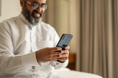 Browsing social media, an Arab Gulf Saudi man wearing traditional attire and a white cap sits on the bed holding a mobile phone, chatting and communicating with family and friends, using a modern and advanced technological device, working remotely and completing the required tasks.