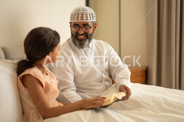 رجل عربي خليجي سعودي يرتدي الثوب التقليدي والقبعة البيضاء يجلس على السرير يقوم بقراءة القصص المسليه لابنته، مفهوم الترابط الأسري، مشاركة قراءه الروايات المفيده مع الأبناء قبل النوم، الشعور بالأمان والحب مع العائله