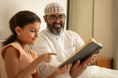 مفهوم الترابط الأسري، مشاركة قراءه الروايات المفيده مع الأبناء قبل النوم، رجل عربي خليجي سعودي يرتدي الثوب التقليدي والقبعة البيضاء يجلس على السرير يقوم بقراءة القصص المسليه لابنته، الشعور بالأمان والحب مع العائله