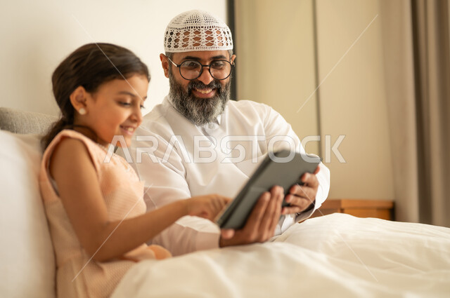 Expressions of happiness and joy, a Saudi Arabian Gulf girl wearing a dress sitting on the bed with her father using a tablet, watching recreational and educational activities, following available online educational platforms, integrating learning with technology and tech.