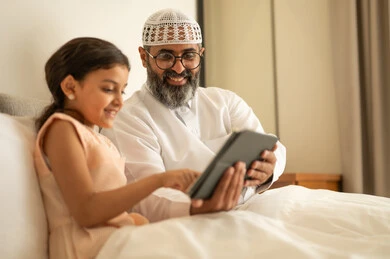 ايماءات السعادة و السرور، فتاة عربية خليجية سعودية ترتدي فستان تجلس على السرير برفقة والدها وتستخدم الجهاز اللوحي، مشاهدة الأنشطة الترفيهية والتعليمية، متابعة منصات التعليم الالكتروني المتاحة عبر الانترنت، دمج التعلم مع التقنية والتكنولوجيا