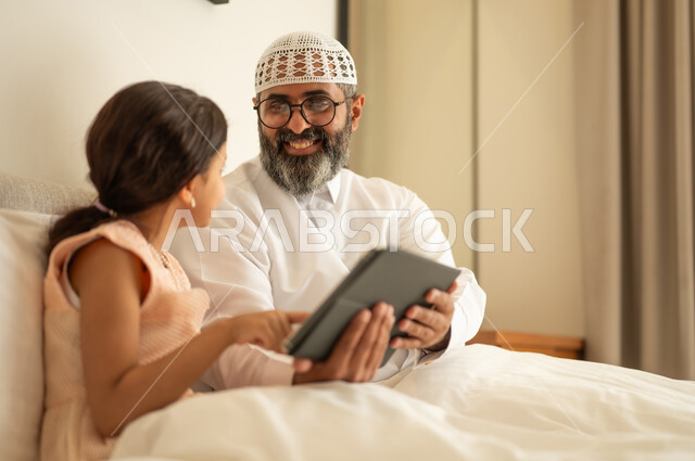 فتاة عربية خليجية سعودية ترتدي فستان تجلس على السرير برفقة والدها وتستخدم الجهاز اللوحي، مشاهدة الأنشطة الترفيهية والتعليمية، متابعة منصات التعليم الالكتروني المتاحة عبر الانترنت، دمج التعلم مع التقنية والتكنولوجيا، ايماءات السعادة و السرور