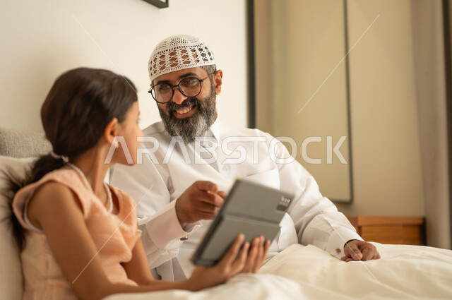 متابعة منصات التعليم الالكتروني المتاحة عبر الانترنت، دمج التعلم مع التقنية والتكنولوجيا، مشاهدة الأنشطة الترفيهية والتعليمية، فتاة عربية خليجية سعودية ترتدي فستان تجلس على السرير برفقة والدها وتستخدم الجهاز اللوحي، ايماءات السعادة و السرور